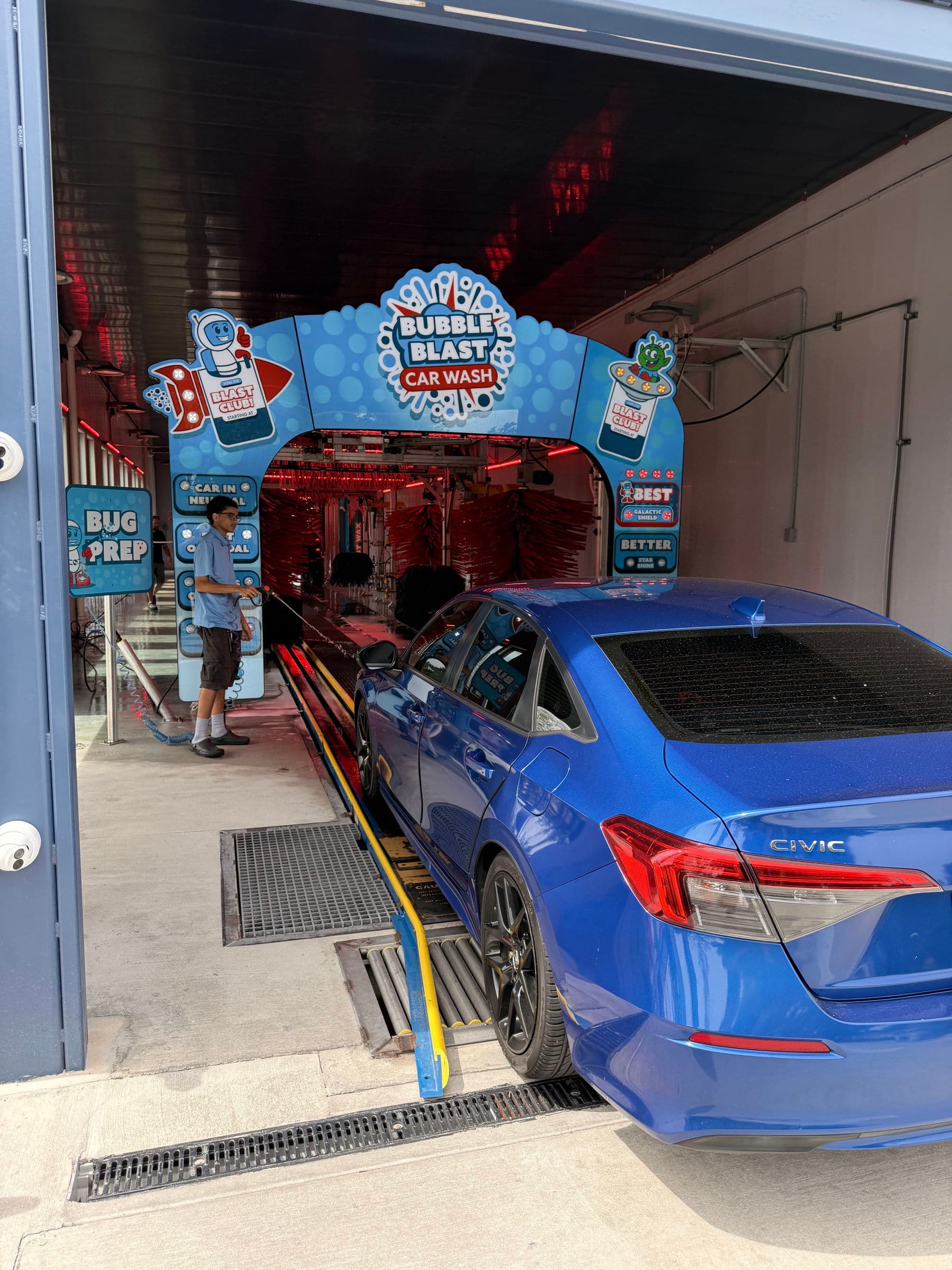 A blue sedan pulling into the Bubble Blast tunnel under the branded entrance arch while an attendant preps it
