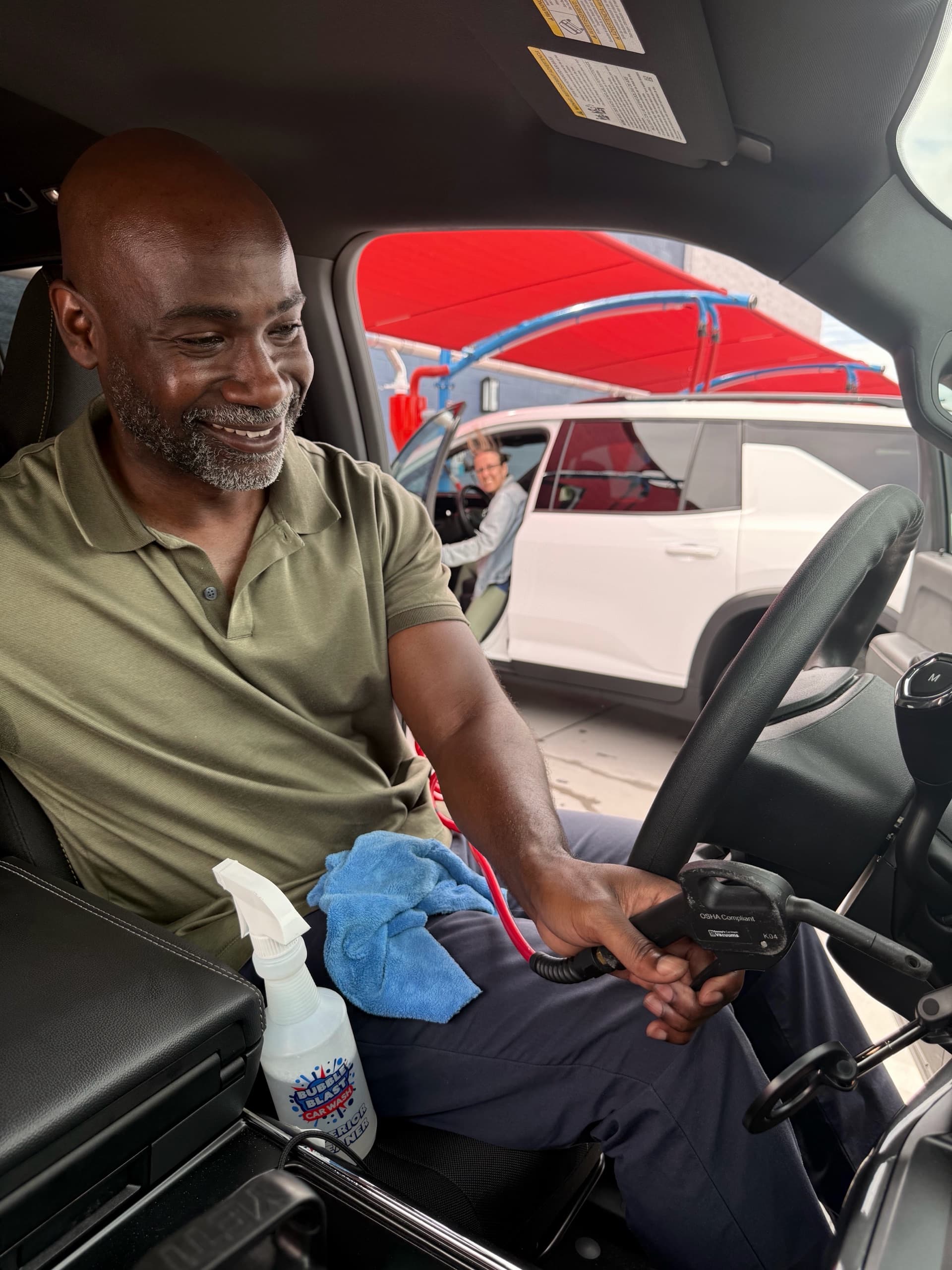 A smiling customer using a free vacuum from the driver's seat, with a Bubble Blast interior cleaner bottle on the console